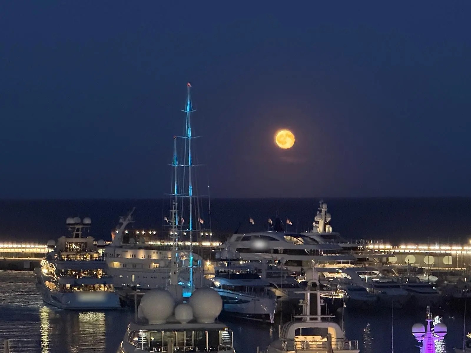 Yachts lit up in the harbour at night, a tall mast lit in blue and the full moon rising over the sea.